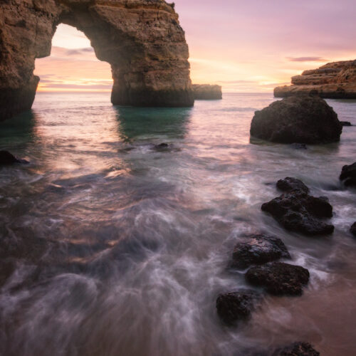 Arco de roca natural en el Algarve con aguas turquesas al atardecer, rodeado de formaciones rocosas y olas suaves