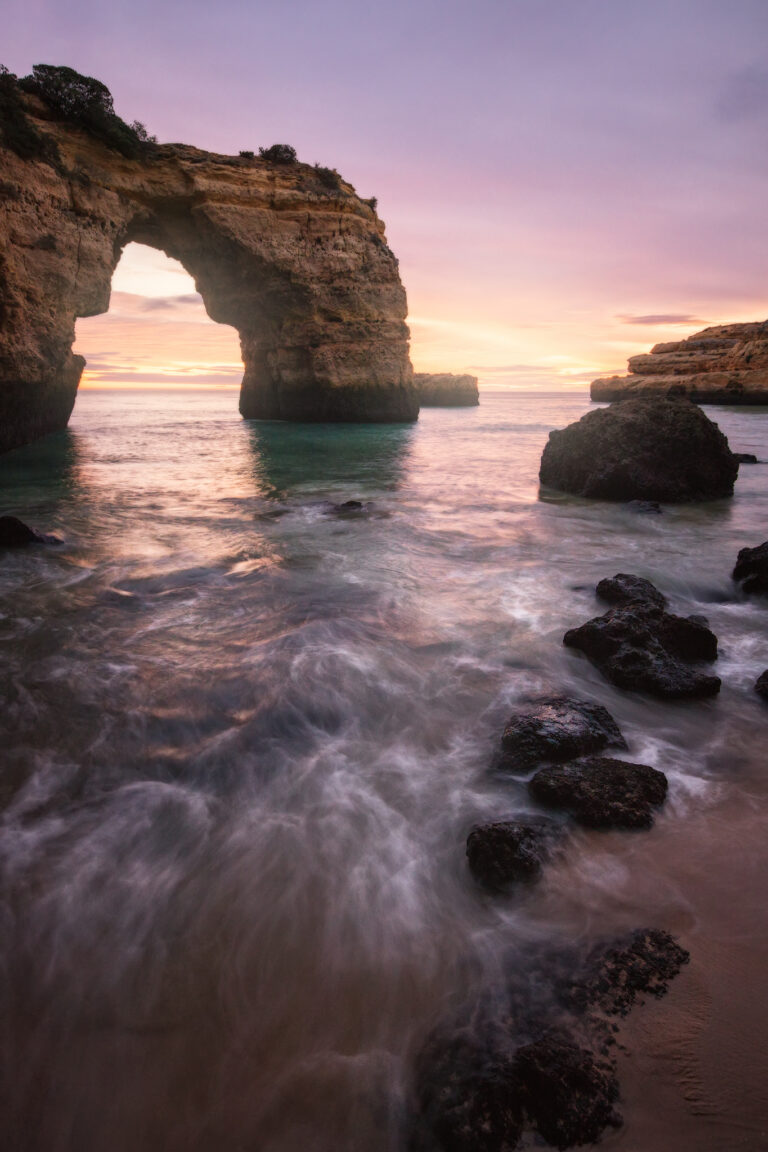 Arco de roca natural en el Algarve con aguas turquesas al atardecer, rodeado de formaciones rocosas y olas suaves