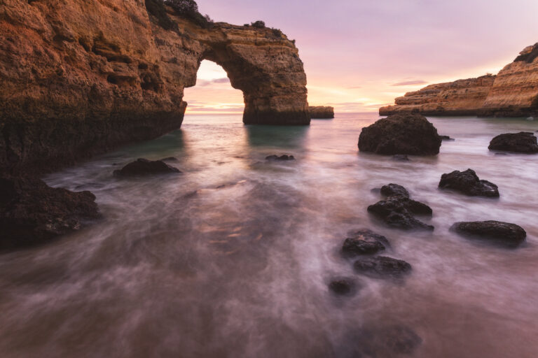 Arco de roca natural en acantilado costero al atardecer, rodeado de agua turquesa y rocas oscuras