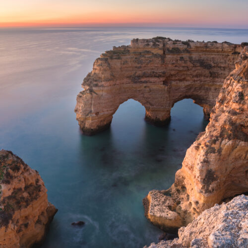 Formación rocosa con dos arcos naturales en acantilados del Algarve al atardecer, junto a islotes de piedra en aguas cristalinas