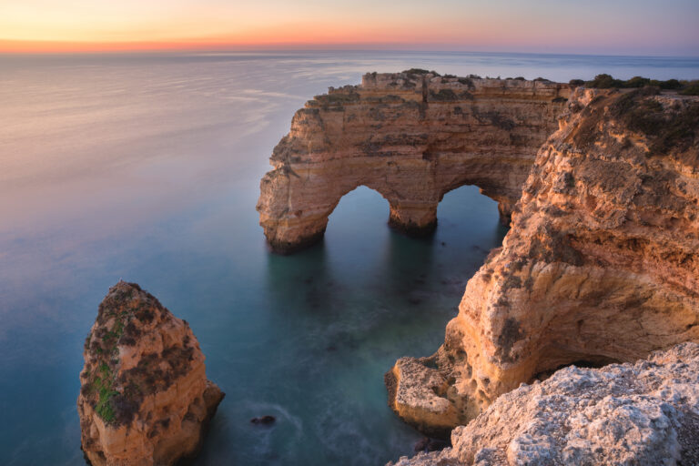 Formación rocosa con dos arcos naturales en acantilados del Algarve al atardecer, junto a islotes de piedra en aguas cristalinas