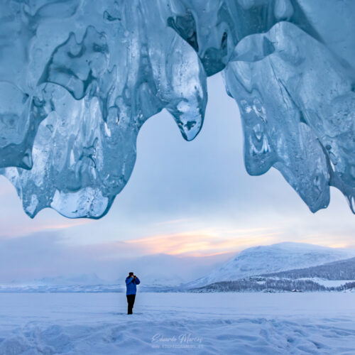 Persona bajo arco de hielo en lago congelado con montañas nevadas al atardecer en Abisko