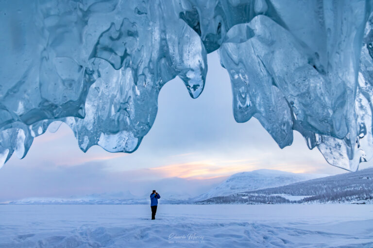 Persona bajo arco de hielo en lago congelado con montañas nevadas al atardecer en Abisko