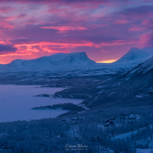 Atardecer alpino sobre lago helado rodeado de montañas nevadas y bosques de coníferas en Abisko