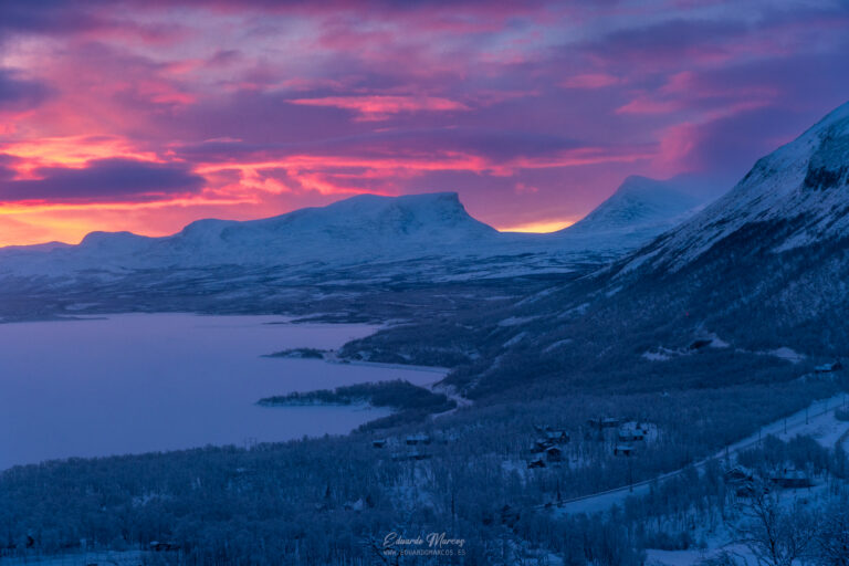 Atardecer alpino sobre lago helado rodeado de montañas nevadas y bosques de coníferas en Abisko