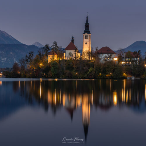 Iglesia de la Asunción en islote del lago Bled al atardecer, reflejada en aguas tranquilas con montañas nevadas de fondo