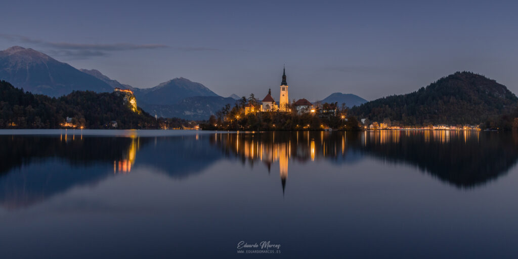 Iglesia de la Asunción reflejada en las aguas tranquilas del lago Bled al atardecer, rodeada de bosques y montañas nevadas