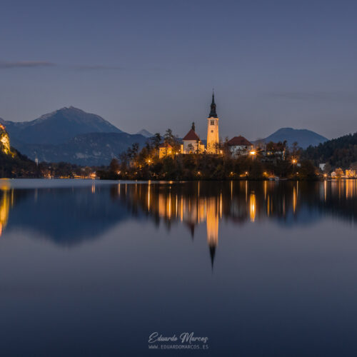Iglesia de la Asunción reflejada en las aguas tranquilas del lago Bled al atardecer, rodeada de bosques y montañas nevadas