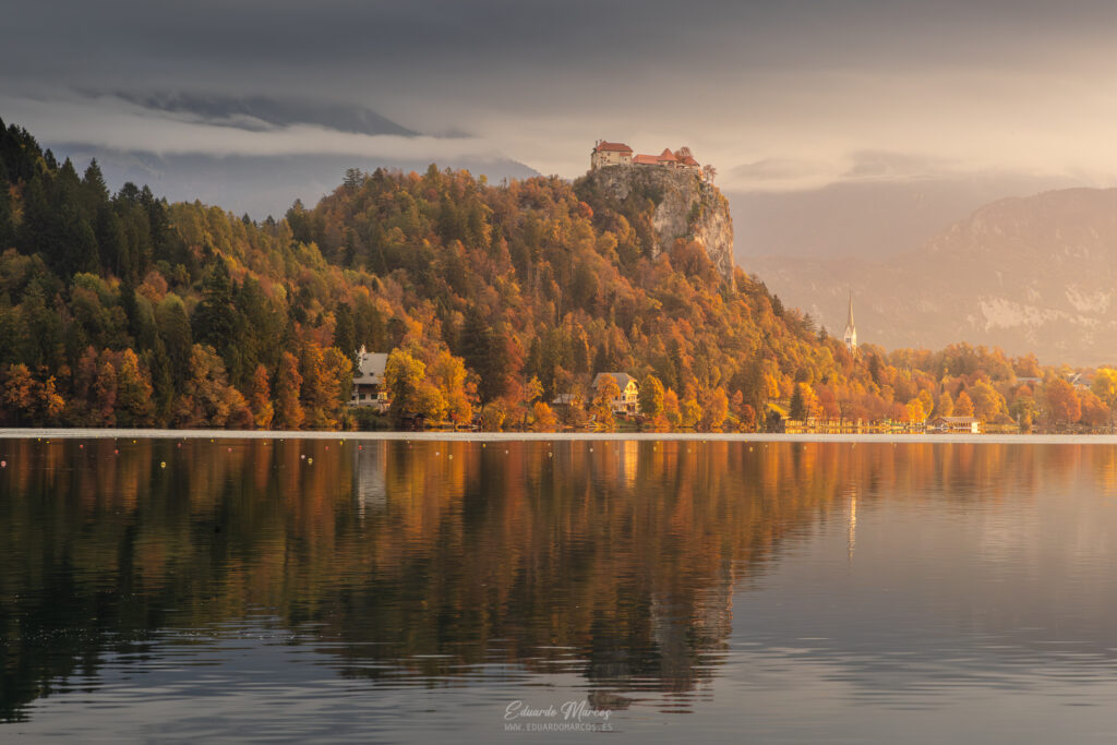 Lago Bled en otoño con castillo en acantilado, bosque dorado reflejado en agua tranquila y montañas al fondo