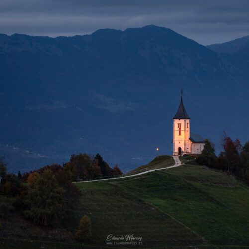 Iglesia de San Jorge iluminada al atardecer en colina verde con montañas azules de fondo, Eslovenia