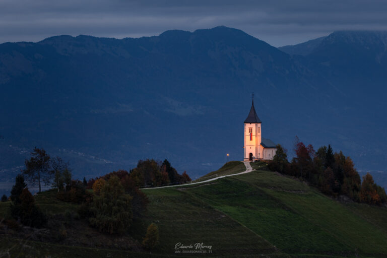 Iglesia de San Jorge iluminada al atardecer en colina verde con montañas azules de fondo, Eslovenia