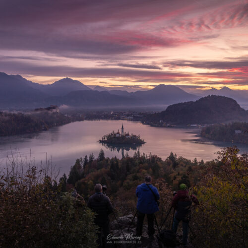 Tres senderistas contemplando el atardecer sobre el lago Bled con la iglesia en la isla y montañas al fondo