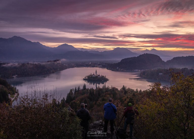 Tres senderistas contemplando el atardecer sobre el lago Bled con la iglesia en la isla y montañas al fondo