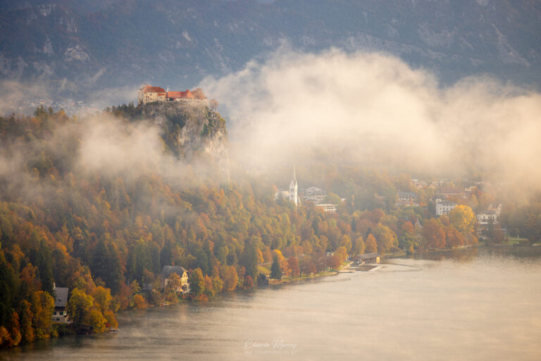 Castillo de Bled sobre acantilado rocoso rodeado de bosque otoñal y niebla, con iglesia y lago al fondo en Eslovenia