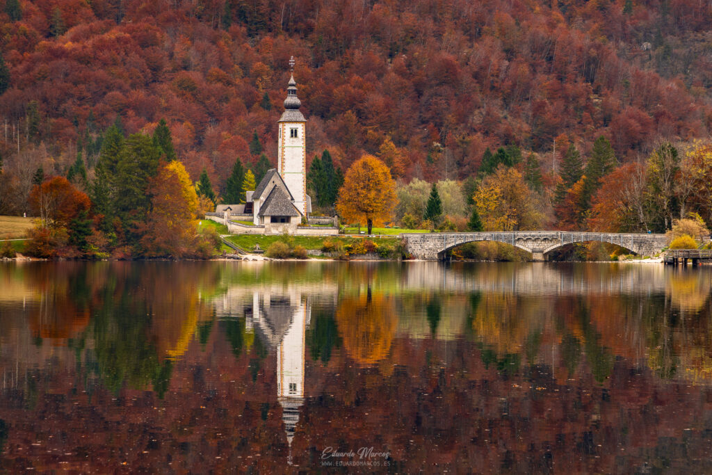 Iglesia de piedra blanca con campanario junto a un lago alpino rodeado de bosque otoñal en tonos rojizos y dorados