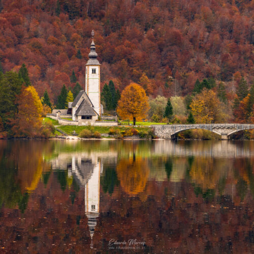 Iglesia de piedra blanca con campanario junto a un lago alpino rodeado de bosque otoñal en tonos rojizos y dorados