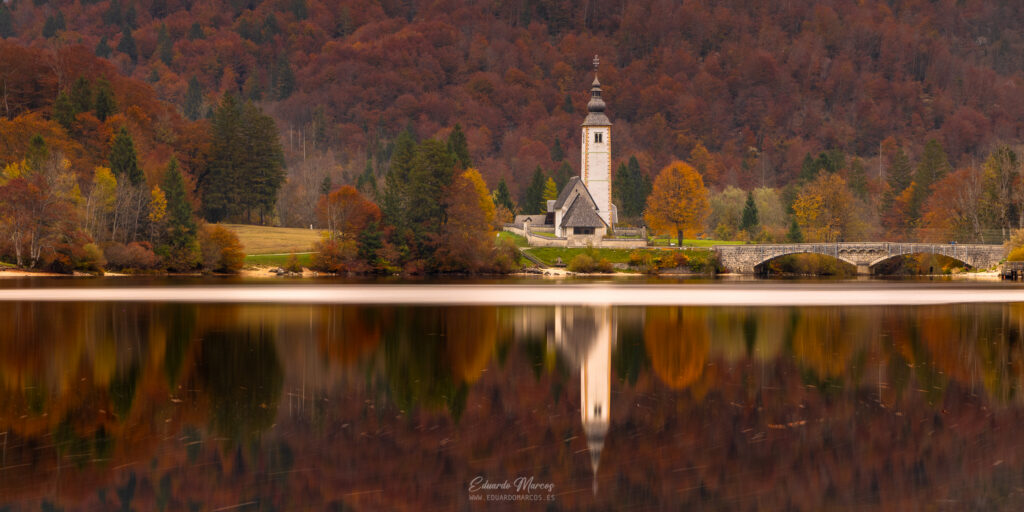 Iglesia de San Juan junto a lago alpino rodeada de bosque otoñal con reflejo espejo en agua tranquila
