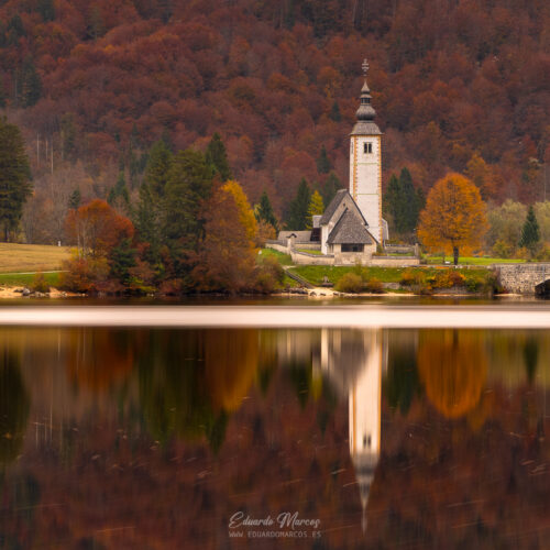 Iglesia de San Juan junto a lago alpino rodeada de bosque otoñal con reflejo espejo en agua tranquila