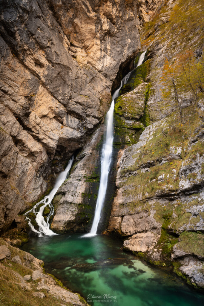 Cascada de agua turquesa cayendo entre acantilados rocosos cubiertos de musgo verde en un cañón estrecho