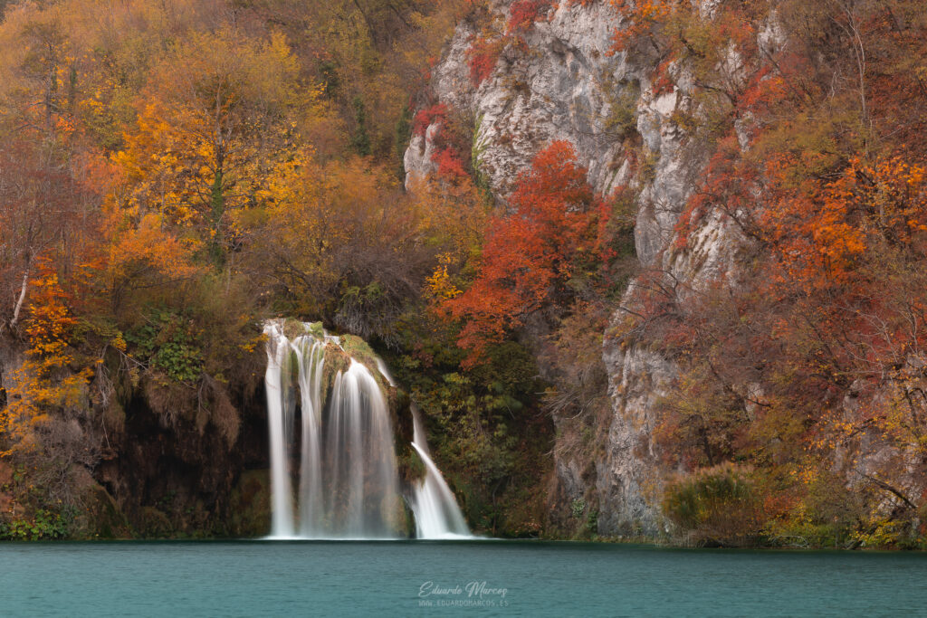 Cascada de agua turquesa entre acantilados rocosos con bosque de otoño en tonos naranjas y rojos