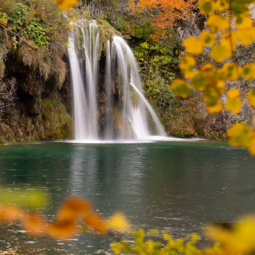 Cascada con piscina turquesa rodeada de rocas musgosas y vegetación otoñal amarilla y anaranjada