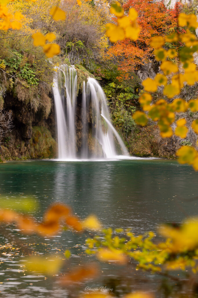 Cascada con piscina turquesa rodeada de rocas musgosas y vegetación otoñal amarilla y anaranjada