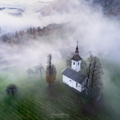Iglesia blanca con campanario entre niebla y montañas boscosas en valle alpino esloveno