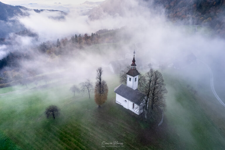 Iglesia blanca con campanario entre niebla y montañas boscosas en valle alpino esloveno