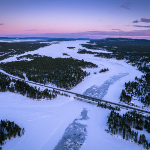 Río congelado rodeado de bosque boreal al atardecer, con carretera atravesando el paisaje nevado de Abisko