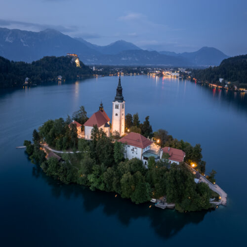 Iglesia de la Asunción en isla de Bled rodeada de bosque, lago azul y montañas al atardecer