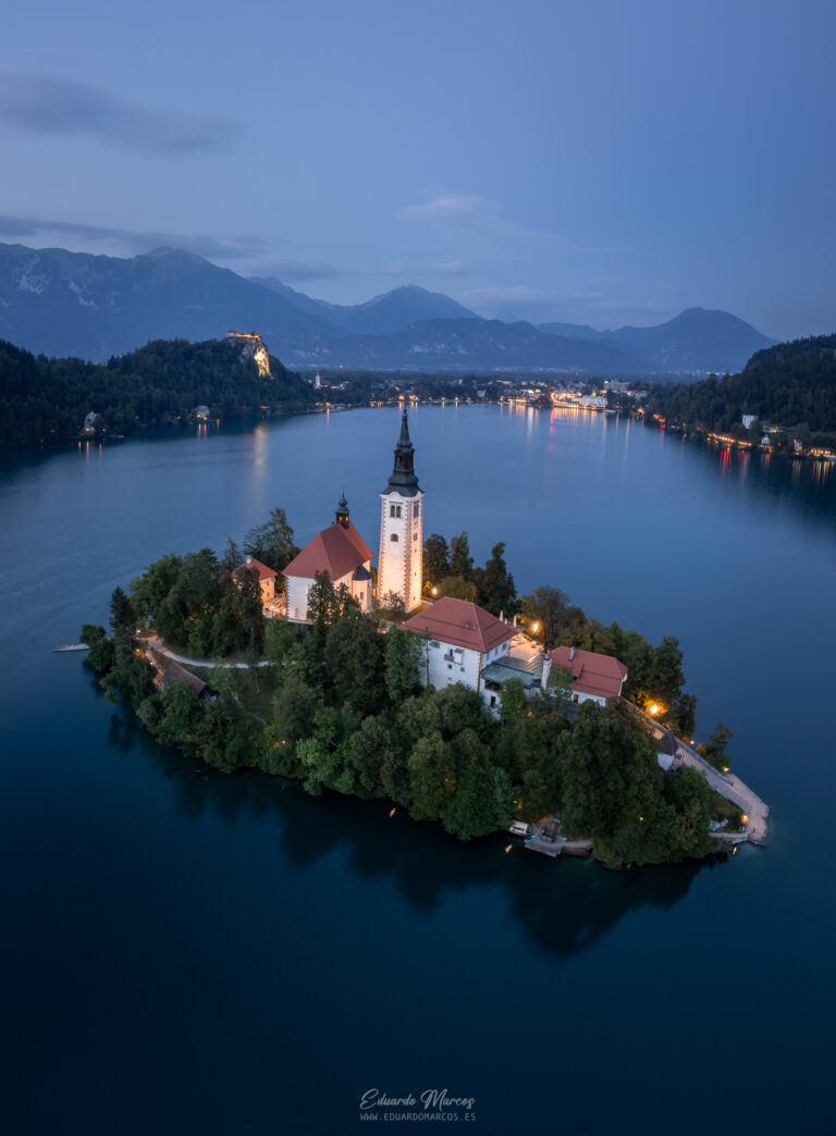 Iglesia de la Asunción en isla de Bled rodeada de bosque, lago azul y montañas al atardecer