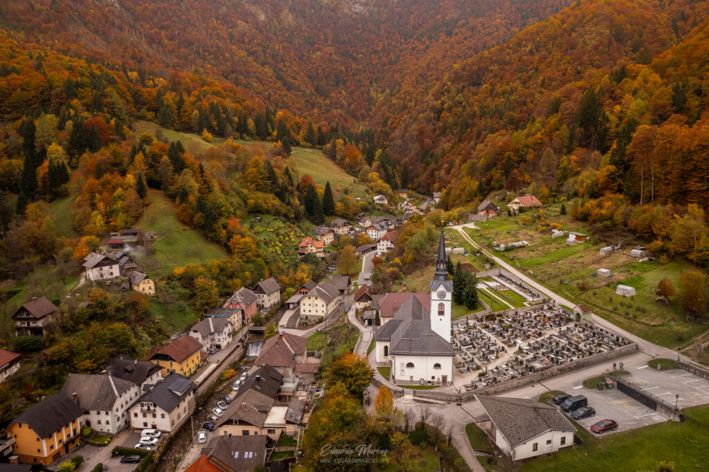 Valle alpino con pueblo, iglesia blanca y bosque otoñal en montañas densamente arboladas