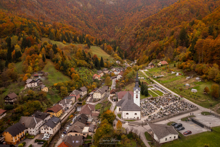 Valle alpino con pueblo, iglesia blanca y bosque otoñal en montañas densamente arboladas