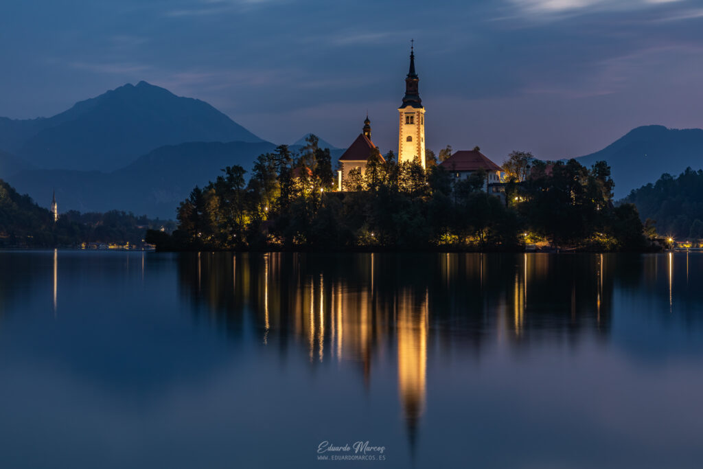 Iglesia de la Asunción en islote del lago Bled al atardecer, reflejada en aguas tranquilas con montañas de fondo