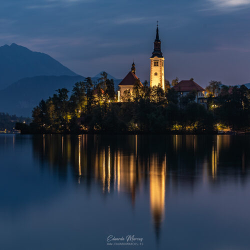 Iglesia de la Asunción en islote del lago Bled al atardecer, reflejada en aguas tranquilas con montañas de fondo