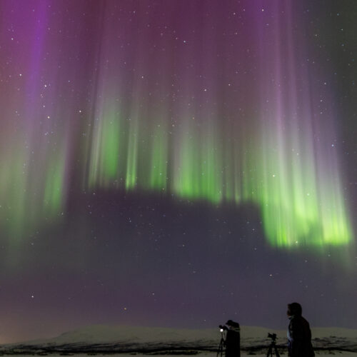 Aurora boreal verde y púrpura sobre paisaje nevado con dos fotógrafos y trípodes en Abisko