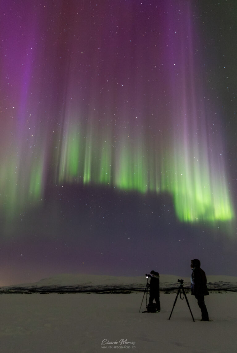 Aurora boreal verde y púrpura sobre paisaje nevado con dos fotógrafos y trípodes en Abisko