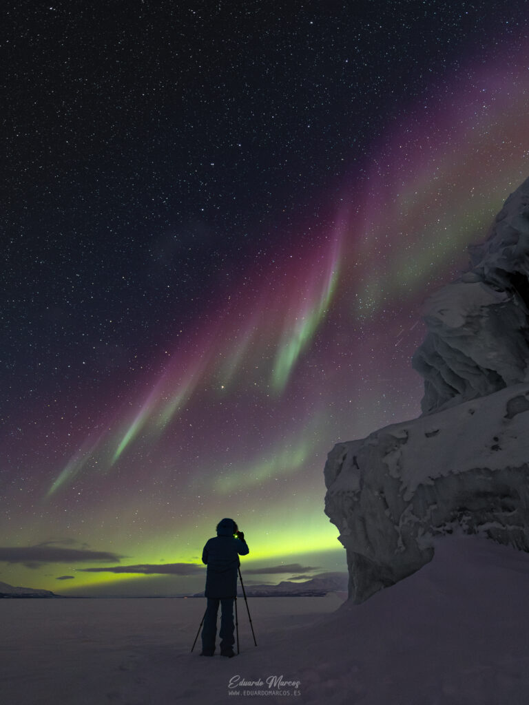 Fotógrafo con trípode bajo aurora boreal verde y rosa sobre hielo y nieve en Abisko, Laponia
