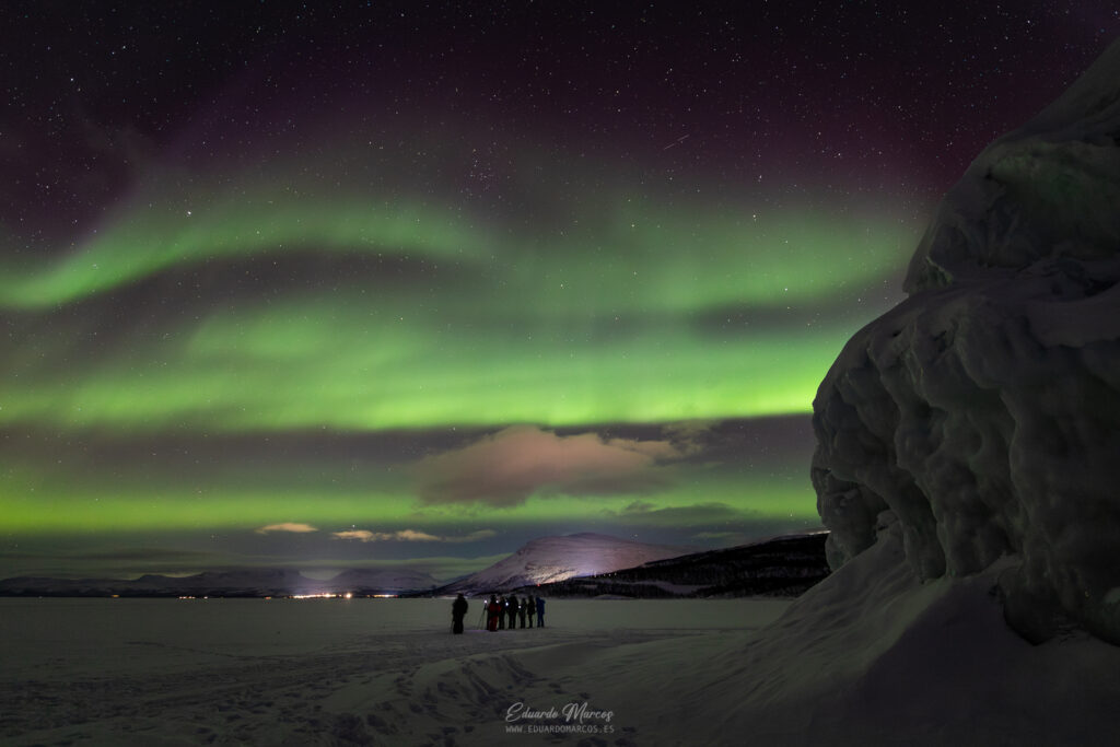 Aurora boreal verde ondulante sobre lago congelado con grupo de personas y rocas en primer plano, Abisko