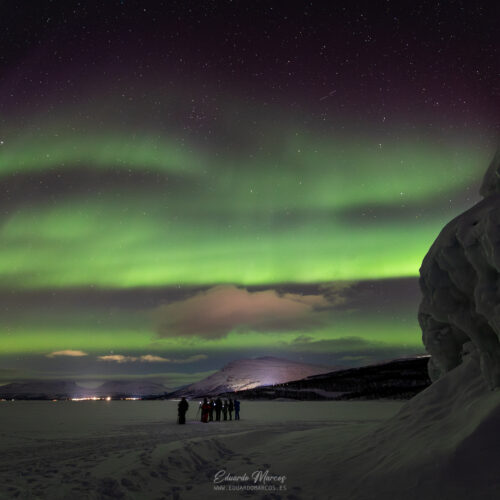 Aurora boreal verde ondulante sobre lago congelado con grupo de personas y rocas en primer plano, Abisko
