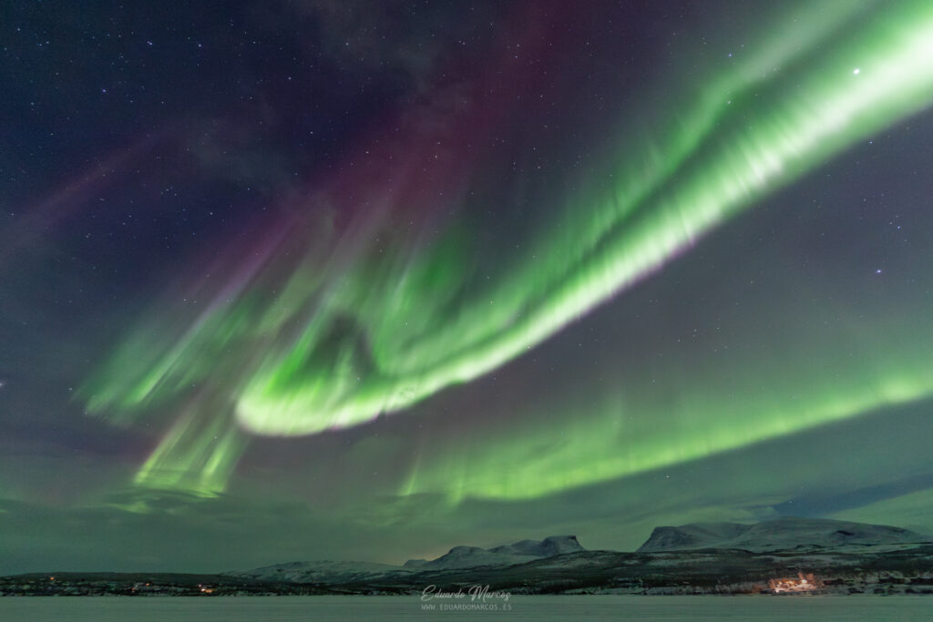 Aurora boreal verde intenso ondulante sobre montañas nevadas y lago congelado en la noche estrellada de Abisko