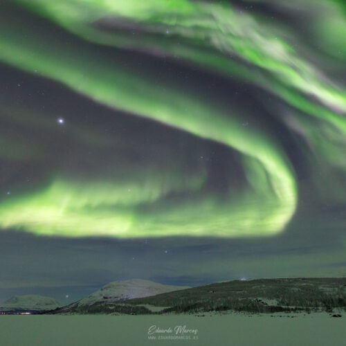 Aurora boreal verde intenso ondulante sobre paisaje nevado de Abisko con montaña al fondo y cielo estrellado