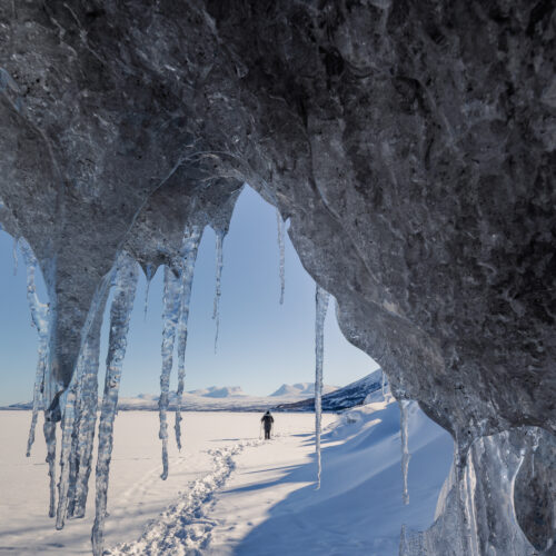 Cueva de hielo con estalactitas en el lago congelado de Abisko, Suecia. Persona en el fondo caminando sobre nieve con montaña al horizonte.