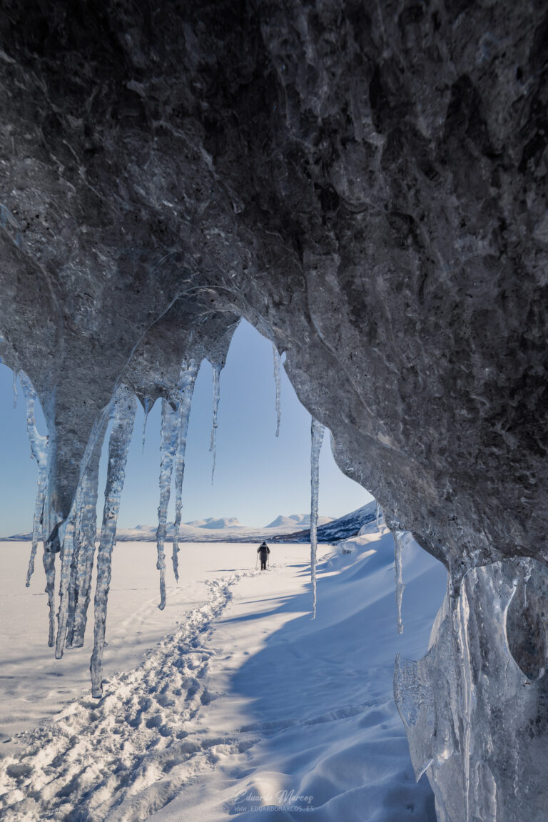 Cueva de hielo con estalactitas en el lago congelado de Abisko, Suecia. Persona en el fondo caminando sobre nieve con montaña al horizonte.